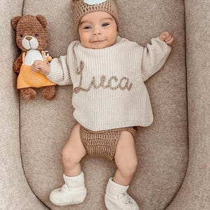 以下が含まれることがあります： 前面に「Luca」と刺繍されたベージュのニットセーター、お揃いのオムツカバー、クマの帽子を着た赤ちゃん。赤ちゃんは白いニットのブーティーも履いています。オレンジ色のドレスを着た茶色のテディベアのおもちゃが左側にあります。
