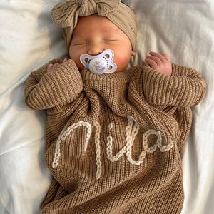 Peut inclure: Un bébé endormi portant un pull en tricot marron avec le nom "Mila" brodé en blanc. Le bébé porte également un bandeau assorti avec un nœud et une sucette.