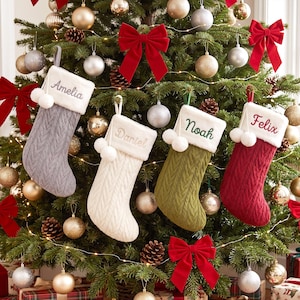 Peut inclure: Quatre chaussettes de Noël personnalisées en tricot torsadé gris, blanc, vert olive et rouge. Chaque chaussette présente un revers en fausse fourrure blanche avec deux pompons blancs et des prénoms brodés : Amelia, Daniel, Noah et Felix.