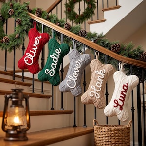 May include: Five bone-shaped Christmas stockings in red, green, gray, tan, and white, each featuring a cable-knit texture and a personalized name in script lettering: Oliver, Sadie, Clover, Hank, and Luna, hanging from a wooden staircase railing.
