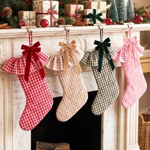 Puede incluir: Cuatro calcetines navideños personalizados colgados de una repisa de chimenea. Cada calcetín presenta un patrón de cuadros vichy en rojo, beige, verde oscuro o rosa, un puño con volantes y un lazo de terciopelo. Los nombres Daniel, Amelia, Noah y Lucy están bordados.
