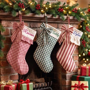 Peut inclure: Trois chaussettes de Noël suspendues. La première est à carreaux rouges et blancs avec un nœud rouge et le nom Rowan. La deuxième est à carreaux verts et blancs avec un nœud vert et le nom Teddy. La troisième est à rayures rouges et blanches avec un nœud et le nom Dylan.