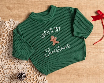 Op de afbeelding: Een groene gebreide kersttrui met de tekst "LUCA'S 1ST Christmas" en een gingerbread man-afbeelding. De trui is gemaakt van een geribbelde gebreide stof. Een rood lint is zichtbaar op de achtergrond.