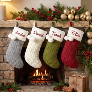 Peut inclure: Quatre chaussettes de Noël en tricot torsadé gris, blanc, vert olive et rouge. Chaque chaussette présente un revers en fausse fourrure blanche avec deux pompons blancs et les prénoms brodés Amelia, Daniel, Noah et Felix en rouge.