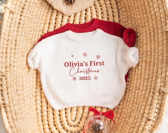 Op de afbeelding: Een witte en rode baby trui met de tekst "Olivia's First Christmas 2025" in het rood. De trui is opgevouwen op een gevlochten mand. Een transparante ornament met een dennenappel en rode strik staat op de voorgrond.