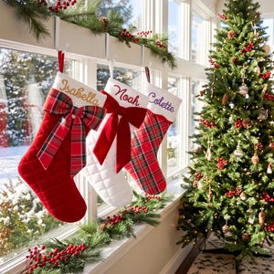 Peut inclure: Trois chaussettes de Noël personnalisées suspendues à des crochets blancs. La première est rouge avec un nœud écossais et le nom Isabella. La deuxième est blanche avec un nœud en velours rouge et le nom Noah. La troisième est écossaise avec le nom Colette.