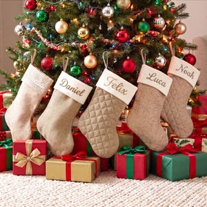 May include: Five beige Christmas stockings with white cuffs hanging in a row. Each cuff is embroidered with a name: Amelia, Daniel, Felix, Luca, and Noah. The stockings have a quilted texture and are displayed in front of a decorated tree.