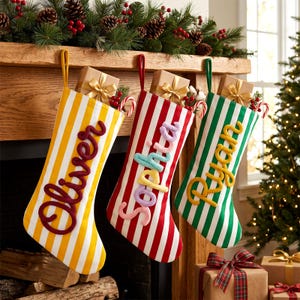 Pode incluir: Três meias de Natal penduradas com listras verticais em amarelo, vermelho e verde. Cada meia é personalizada com um nome tricotado em uma cor contrastante: Oliver em vermelho escuro, Sophia em fio multicolorido e Ryan em amarelo.