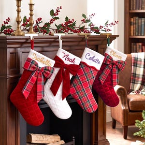 Peut inclure: Quatre chaussettes de Noël suspendues à une cheminée en bois. La première est en velours rouge avec un nœud écossais et le nom Isabella. La deuxième est en tissu blanc matelassé avec un nœud rouge et le nom Noah. La troisième est à carreaux rouges avec le nom Colette. La quatrième est en velours rouge avec un nœud écossais et le nom Oliver.