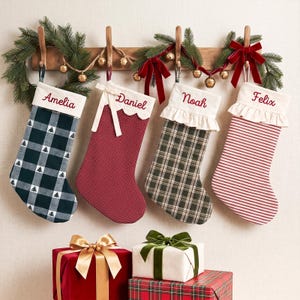 Peut inclure: Quatre chaussettes de Noël personnalisées suspendues à un support en bois avec de la verdure et des clochettes. Les chaussettes portent les noms Amelia, Daniel, Noah et Felix brodés en rouge. Elles présentent des motifs à carreaux et rayures.