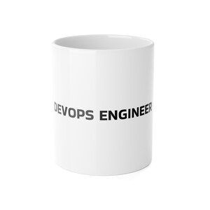 May include: A white ceramic mug featuring the text DEVOPS ENGINEER printed in bold black capital letters across the center. The mug has a smooth, cylindrical body with a clean, minimalist design and a glossy finish, standing upright against a white background.