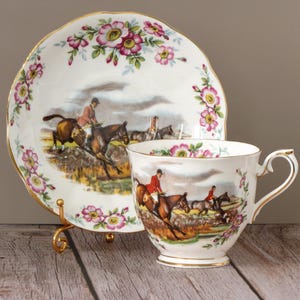 May include: A vintage teacup and saucer set. The white porcelain features a hunting scene with riders on horseback, and is accented with pink floral designs and gold trim. The saucer is scalloped.