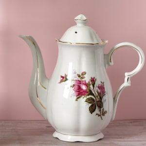 May include: White porcelain teapot with a lid, spout, and handle. The teapot is decorated with pink and red roses and green leaves. Gold trim accents the edges and handle. The teapot is on a wooden surface against a pink background.