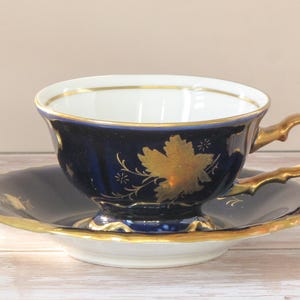 May include: A vintage teacup and saucer set. The teacup and saucer are navy blue with gold trim. A gold leaf design decorates the teacup. The handle and rim of the teacup are also gold. The saucer has a scalloped edge.