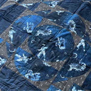 May include: A quilted blanket with a space theme. The design features astronauts floating in space, with a dark blue and gray color scheme. The quilt is made of fabric squares and has a stitched pattern.