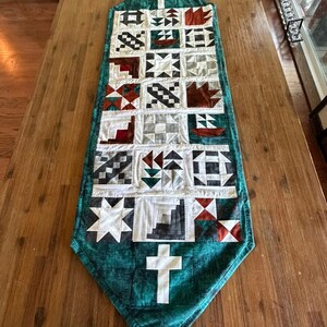 May include: A quilted table runner with a teal green border and a patchwork design. The patchwork includes squares with various patterns in white, black, red, and gray. White crosses are at the top and bottom.