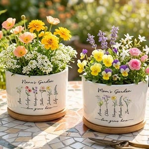 Könnte beinhalten: Zwei weiße Keramik-Blumentöpfe mit Blumenarrangements stehen auf einem Mosaiktisch. Auf einem Topf steht "Nana's Garden" und auf dem anderen "Mom's Garden". Die Töpfe stehen auf Holzuntersetzern. Der Text "love grows here" steht auf den Töpfen.