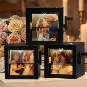Könnte beinhalten: Drei schwarze Holz-Fotowürfel mit beleuchteten Bildern von Menschen. Jeder Würfel hat eine Kurbel an der Seite. Der Hintergrund umfasst einen Rosenstrauß und brennende Kerzen, die eine warme, sentimentale Atmosphäre schaffen.