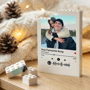 Könnte beinhalten: Ein weißer, aus Ziegeln gebauter Rahmen mit einem Foto eines Paares, dem Text "Your Favourite Song" und einer Musikplayer-Oberfläche. Der Rahmen steht auf einer Holzoberfläche, mit einer gemütlichen Decke und Tannenzapfen im Hintergrund.