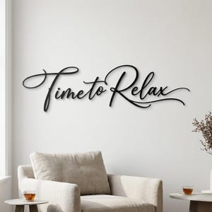 Könnte beinhalten: Schwarze Metall-Wandkunst mit dem geschwungenen Schriftzug „Time to Relax“ an einer weißen Wand. Die Kunst befindet sich über einem beigen Sessel, mit einem kleinen Beistelltisch und einer Tasse Tee.