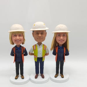 以下が含まれることがあります： 建設作業員のボブルヘッド人形が3体。各フィギュアは白いヘルメット、濃いネイビーのシャツ、オレンジ色の安全ベストを着用しています。1体のフィギュアは黄色の安全ベストを着用しています。フィギュアは白い円形の台座の上に立っています。