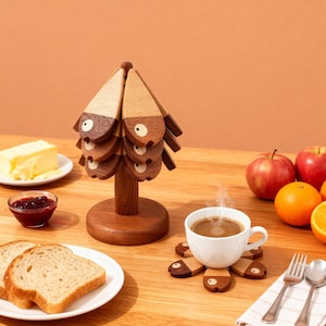 Puede incluir: Un juego de posavasos de madera con forma de árbol con una base a juego. Los posavasos son marrones y beige con un diseño caprichoso de pez. Una taza de café blanca se encuentra sobre un posavasos. Manzanas, naranjas, mantequilla, mermelada y pan también están sobre la mesa.