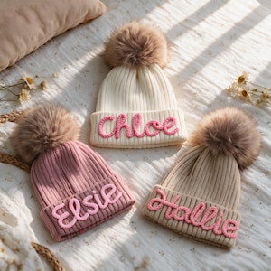 May include: Three ribbed knit beanies with large faux fur pom-poms. The white beanie features the name Chloe in pink script, the pink beanie says Elsie in pink, and the beige beanie displays the name Hallie in pink script.