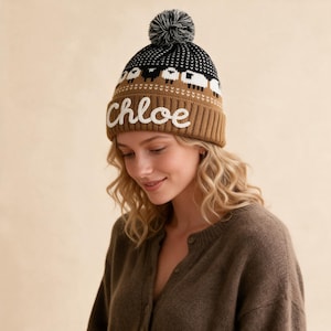 May include: A knit beanie with a brown base and a black top section featuring a white sheep pattern. The cuff displays the name Chloe in white embroidered script lettering. The hat is topped with a black and white pom-pom.