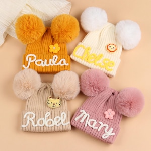 May include: Four knitted beanies in various colours, each with two large pom-poms. The beanies are mustard yellow, cream, beige, and pink, with the names "Paula", "Chelse", "Robel", and "Mary" embroidered on them. Each has a decorative embellishment.