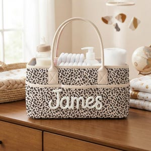 Peut inclure: Un panier de rangement rectangulaire avec un motif léopard marron et crème et deux anses. Le devant du panier affiche le prénom James en lettres cursives blanches. Il contient un biberon, des couches et un flacon blanc.