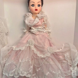 May include: A vintage porcelain doll in a pink and white gown. The doll has a black hair, a veil, and a pearl necklace. The dress features lace trim and decorative embellishments. The doll is a collectible item.