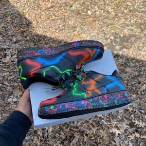 Pu&ograve; includere: Scarpe da ginnastica nere con fulmini al neon rossi, blu e verdi. Le suole hanno un effetto vernice a spruzzo colorato. I lacci sono multicolore.