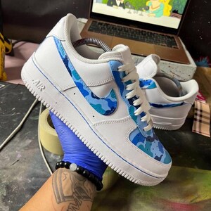 May include: White Air Force 1 sneakers with blue camouflage accents on the Nike swoosh and side panels. The shoes have white laces and a white sole with the word "AIR" visible. The sneakers are held by a person wearing a blue glove.