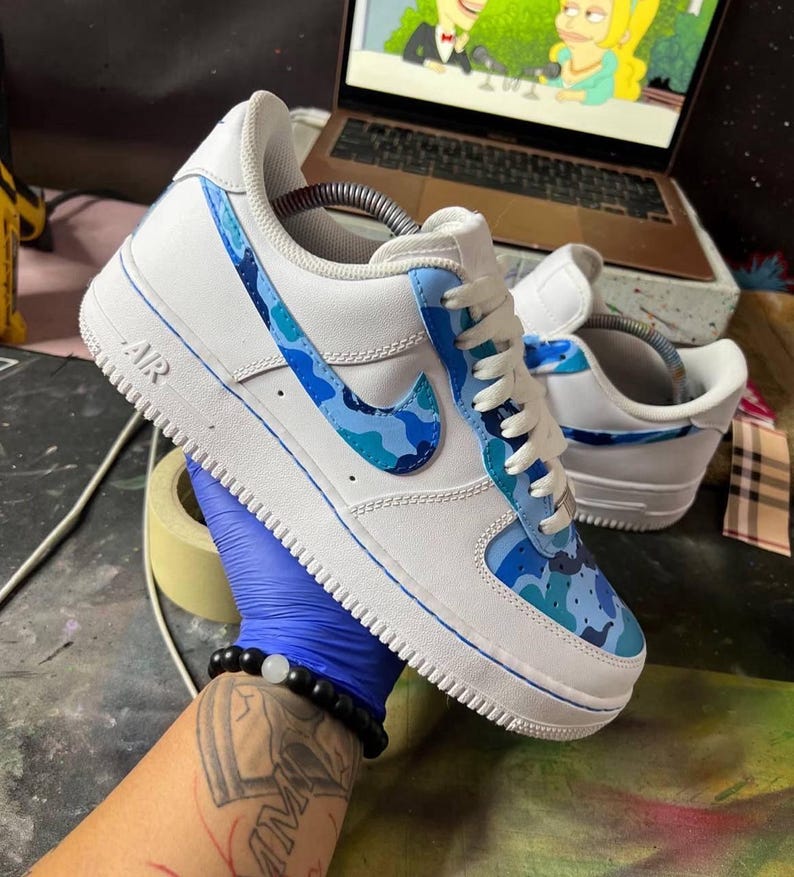 May include: White Air Force 1 sneakers with blue camouflage accents on the Nike swoosh and side panels. The shoes have white laces and a white sole with the word "AIR" visible. The sneakers are held by a person wearing a blue glove.