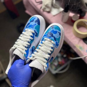 May include: A pair of white sneakers with blue camouflage design on the toe and side panels. The sneakers have white laces and a white sole. The shoes are held by a person wearing a blue glove.