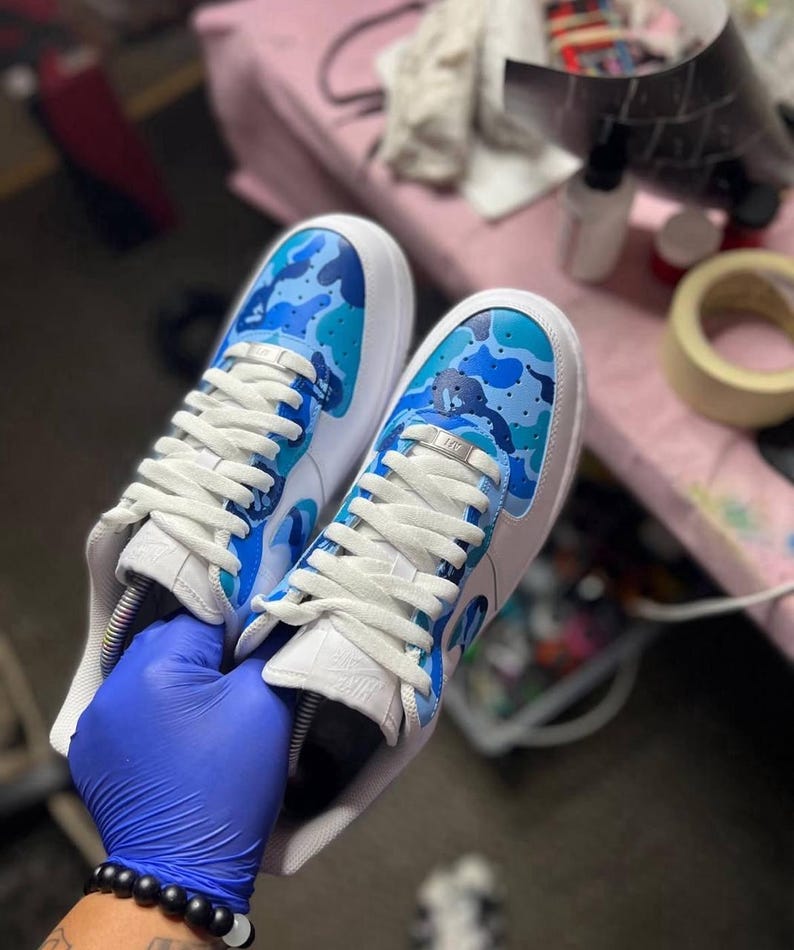 May include: A pair of white sneakers with blue camouflage design on the toe and side panels. The sneakers have white laces and a white sole. The shoes are held by a person wearing a blue glove.