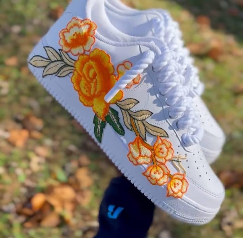 May include: White trainers featuring embroidered orange and yellow flowers and green leaves. The floral design is on the side. The laces are white and thick. The shoes are on a blurred background.