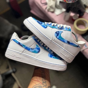 May include: White sneakers with blue camouflage accents. The shoes feature a white base with blue camouflage patterns on the Nike swoosh and heel. The soles and trim are white with a blue accent line. The word "AIR" is visible on the side.