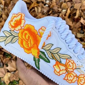 May include: White trainers featuring an embroidered orange and yellow floral design. The flowers and leaves have green and gold stitching. White laces complete the look.