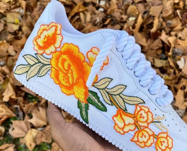 May include: White trainers featuring an embroidered orange and yellow floral design. The flowers and leaves have green and gold stitching. White laces complete the look.