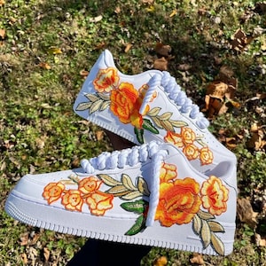 May include: White trainers featuring orange and yellow floral embroidery. The shoes have white laces and a white sole. The floral design includes green leaves and stems, adding colour to the overall design.