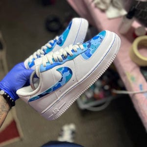 May include: White sneakers with blue camouflage accents on the toe, side, and heel. The sneakers have white laces and a white sole. The word "AIR" is visible on the side of the shoe.