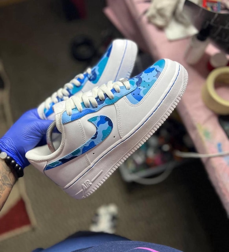 May include: White sneakers with blue camouflage accents on the toe, side, and heel. The sneakers have white laces and a white sole. The word "AIR" is visible on the side of the shoe.