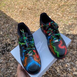 Pu&ograve; includere: Un paio di scarpe da ginnastica Nike Air Force 1 nere con accenti neon colorati in rosso, arancione e blu. I lacci sono multicolori e le scarpe poggiano su una scatola bianca con il logo Nike. Design artistico unico.