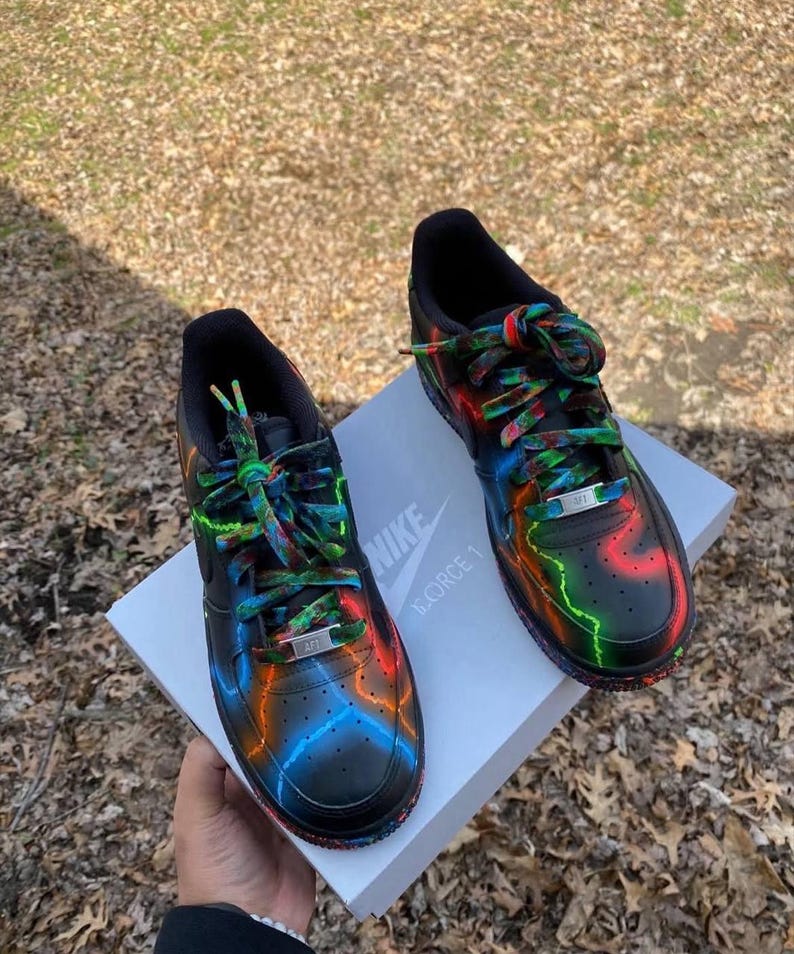 Pu&ograve; includere: Un paio di scarpe da ginnastica Nike Air Force 1 nere con accenti neon colorati in rosso, arancione e blu. I lacci sono multicolori e le scarpe poggiano su una scatola bianca con il logo Nike. Design artistico unico.