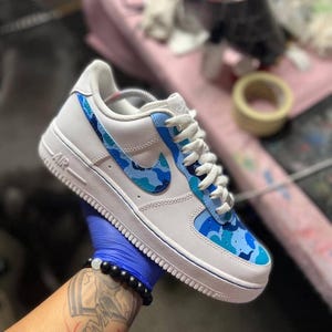 May include: White and blue Air Force 1 sneakers with a custom camouflage design. The shoe features a white base with blue camouflage accents on the Nike swoosh and toe box. White laces and a blue trim complete the design.