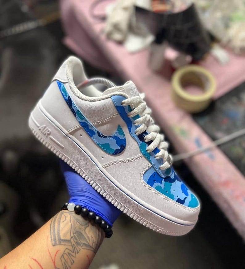 May include: White and blue Air Force 1 sneakers with a custom camouflage design. The shoe features a white base with blue camouflage accents on the Nike swoosh and toe box. White laces and a blue trim complete the design.