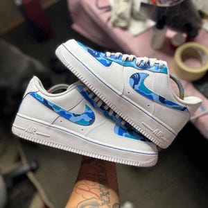 May include: White sneakers with blue camouflage accents. The shoes feature a white base with blue camouflage patterns on the Nike swoosh and heel. The soles and trim are white with a blue accent line. The word "AIR" is visible on the side.