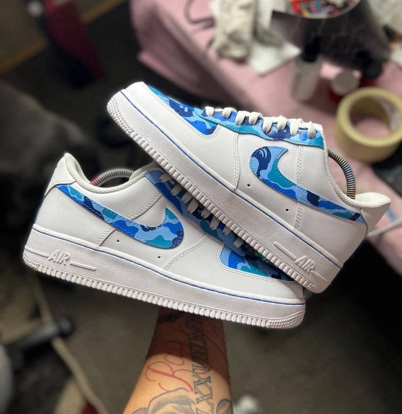 May include: White sneakers with blue camouflage accents. The shoes feature a white base with blue camouflage patterns on the Nike swoosh and heel. The soles and trim are white with a blue accent line. The word "AIR" is visible on the side.