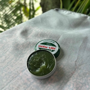 Puede incluir: Lata abierta de bálsamo verde con tapa a juego. El bálsamo es de color verde oscuro y tiene una textura suave similar a un gel. La tapa tiene texto verde y blanco. La lata está sobre una tela estampada en rosa claro y blanco.